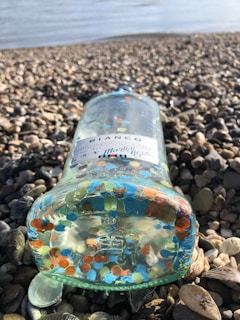 A glass bottle with a label reading 'Bianco Martini Rossi' lies on a pebbled beach. The bottle contains numerous colorful plastic discs and is positioned near the water's edge.