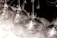 A close-up view of several upside-down wine glasses arranged in neat rows, casting soft reflections and light refractions on a white surface. The glasses have a clear and glossy appearance, highlighting their transparent nature.