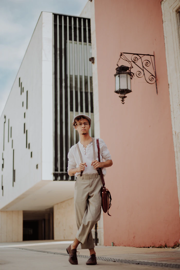 Man in stone pleated trousers and braces in front of a pink wall
