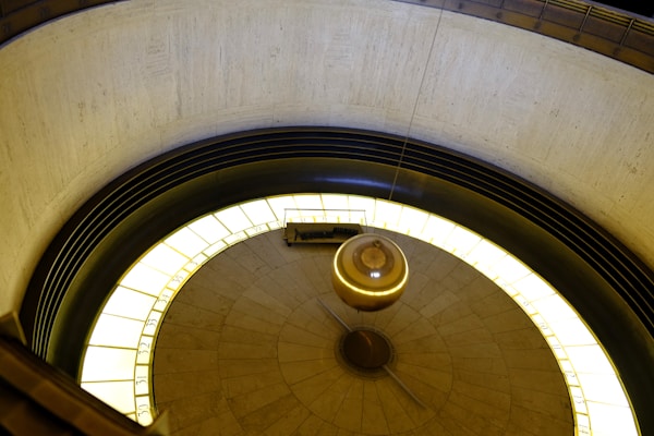 A large, circular pendulum swings over a round platform marked with numbers. The pendulum is suspended from above, and the entire scene is encircled by a strip of light that illuminates the numbers on the perimeter. The architectural design features a blend of stone and metal elements, creating an intriguing visual effect.