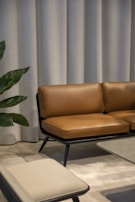 A modern interior with a brown leather sofa set against a backdrop of light grey curtains. Adjacent to the sofa is a low coffee table with a smooth surface and a beige chair in the foreground. A green plant with broad leaves peeks into the scene from the side.