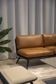 A modern interior with a brown leather sofa set against a backdrop of light grey curtains. Adjacent to the sofa is a low coffee table with a smooth surface and a beige chair in the foreground. A green plant with broad leaves peeks into the scene from the side.