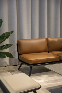 A modern interior with a brown leather sofa set against a backdrop of light grey curtains. Adjacent to the sofa is a low coffee table with a smooth surface and a beige chair in the foreground. A green plant with broad leaves peeks into the scene from the side.