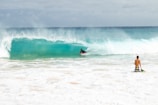 A surfer rides a turquoise wave while another person stands near the shore holding a surfboard. The ocean is vibrant and energetic, with white foam spreading across the beach.