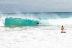 A surfer rides a turquoise wave while another person stands near the shore holding a surfboard. The ocean is vibrant and energetic, with white foam spreading across the beach.