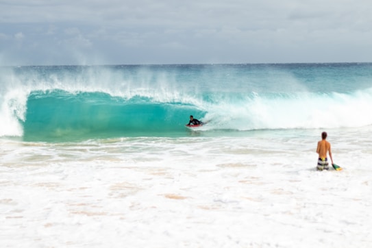 A surfer rides a turquoise wave while another person stands near the shore holding a surfboard. The ocean is vibrant and energetic, with white foam spreading across the beach.