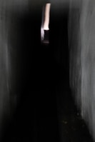 A dark, narrow corridor with dim lighting leads to a small, bright opening at the far end. The walls are textured with a rough surface, and the lack of detail adds a mysterious, unsettling atmosphere. The bright opening provides a stark contrast to the surrounding darkness, drawing attention down the hallway.