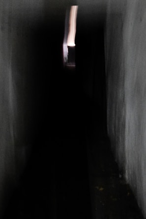 A dark, narrow corridor with dim lighting leads to a small, bright opening at the far end. The walls are textured with a rough surface, and the lack of detail adds a mysterious, unsettling atmosphere. The bright opening provides a stark contrast to the surrounding darkness, drawing attention down the hallway.