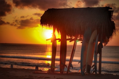 A thatched roof hut with surfboards standing upright underneath it, positioned on a beach with the sun setting over the ocean in the background. The sky is filled with clouds partially illuminated by the setting sun.