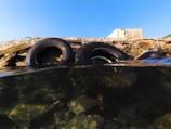 Two rubber tires are partially submerged in clear water, leaning against a concrete structure. The water is transparent, revealing rocks beneath its surface. The sky above is clear and blue, contrasting with the rough texture of the concrete and tires.