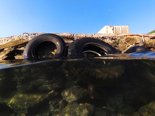 Two rubber tires are partially submerged in clear water, leaning against a concrete structure. The water is transparent, revealing rocks beneath its surface. The sky above is clear and blue, contrasting with the rough texture of the concrete and tires.