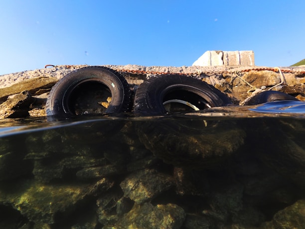 Two rubber tires are partially submerged in clear water, leaning against a concrete structure. The water is transparent, revealing rocks beneath its surface. The sky above is clear and blue, contrasting with the rough texture of the concrete and tires.