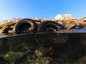 Two rubber tires are partially submerged in clear water, leaning against a concrete structure. The water is transparent, revealing rocks beneath its surface. The sky above is clear and blue, contrasting with the rough texture of the concrete and tires.