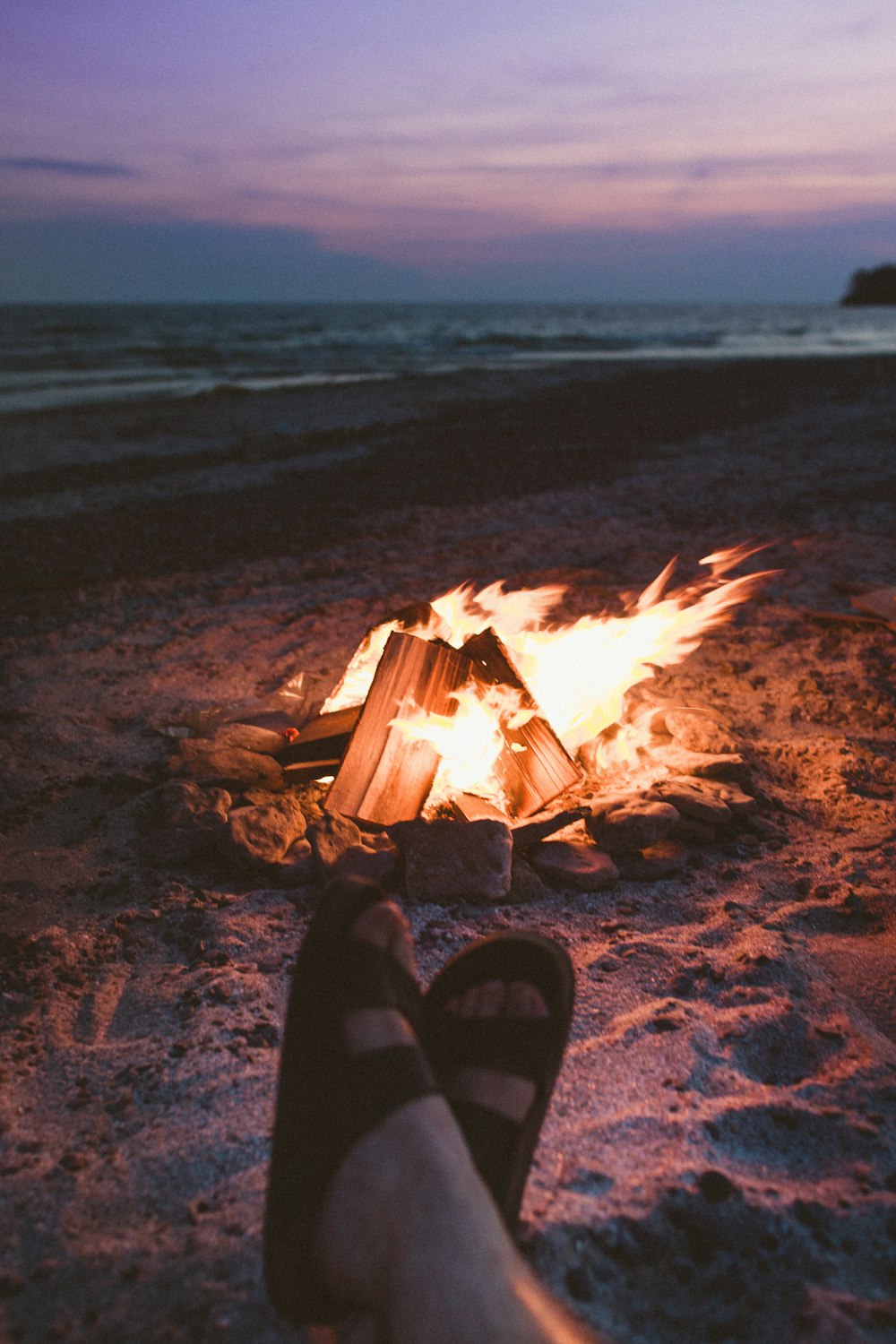 selective focus photography of person wearing black 2-strap slide sandals in front of burned bonfire on beach shore