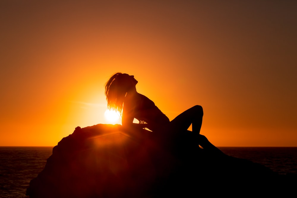 silhouette of woman lying on rock under orange sky during sunset