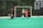 A field hockey game is taking place with a goalkeeper in green and black gear lying on the ground in front of a goalpost. A player in a red uniform is to the right, seemingly moving toward the ball or goal, while another player in a green and white uniform runs alongside. The background shows a scoreboard with championship years listed, and the scene is set on a green artificial turf field.