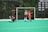 A field hockey game is taking place with a goalkeeper in green and black gear lying on the ground in front of a goalpost. A player in a red uniform is to the right, seemingly moving toward the ball or goal, while another player in a green and white uniform runs alongside. The background shows a scoreboard with championship years listed, and the scene is set on a green artificial turf field.