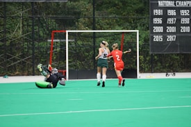 A field hockey game is taking place with a goalkeeper in green and black gear lying on the ground in front of a goalpost. A player in a red uniform is to the right, seemingly moving toward the ball or goal, while another player in a green and white uniform runs alongside. The background shows a scoreboard with championship years listed, and the scene is set on a green artificial turf field.