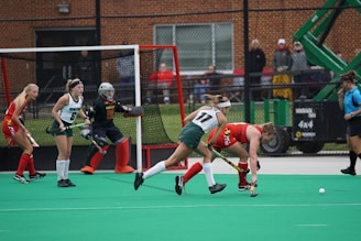 A group of female field hockey players are actively engaged in a game. Two players in green and white uniforms are closely contesting a ball with a player in a red and yellow uniform. A goalkeeper in a black uniform with red accents is positioned in front of the goal. The field is green with a bright red goal and a netted fence. Spectators and an official in a blue top are visible in the background.