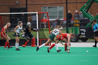 A group of female field hockey players are actively engaged in a game. Two players in green and white uniforms are closely contesting a ball with a player in a red and yellow uniform. A goalkeeper in a black uniform with red accents is positioned in front of the goal. The field is green with a bright red goal and a netted fence. Spectators and an official in a blue top are visible in the background.