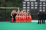 A group of female athletes in red sports uniforms posing for a team photo in front of a banner displaying championship years. They are on a green field with two coaches standing to the side, watching.