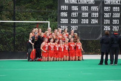 A group of female athletes in red sports uniforms posing for a team photo in front of a banner displaying championship years. They are on a green field with two coaches standing to the side, watching.