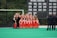 A group of female athletes in red sports uniforms posing for a team photo in front of a banner displaying championship years. They are on a green field with two coaches standing to the side, watching.