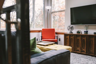 A cozy living room featuring a retro-style wooden cabinet with a modern flat-screen TV mounted above it. A vibrant red armchair sits by large windows that allow natural light to illuminate the space. A yellow cushioned ottoman and a sofa with green pillows add to the comfort. Decorative items, including a vintage fan and a glass vase, are placed on the cabinet. The outside view shows trees with autumn leaves, providing a warm backdrop.
