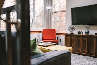 A cozy living room featuring a retro-style wooden cabinet with a modern flat-screen TV mounted above it. A vibrant red armchair sits by large windows that allow natural light to illuminate the space. A yellow cushioned ottoman and a sofa with green pillows add to the comfort. Decorative items, including a vintage fan and a glass vase, are placed on the cabinet. The outside view shows trees with autumn leaves, providing a warm backdrop.