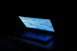 A partially open laptop with a bright screen displaying a blue-toned desktop image, set against a dark background.