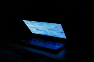 A partially open laptop with a bright screen displaying a blue-toned desktop image, set against a dark background.