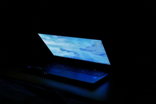 A partially open laptop with a bright screen displaying a blue-toned desktop image, set against a dark background.