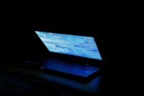 A partially open laptop with a bright screen displaying a blue-toned desktop image, set against a dark background.