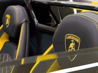 A close-up view of luxury car seats with black leather and yellow stitching, prominently featuring the Lamborghini logo. The headrests are detailed with a gold emblem of a bull surrounded by a shield, indicating the brand identity.