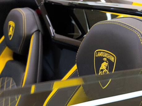 A close-up view of luxury car seats with black leather and yellow stitching, prominently featuring the Lamborghini logo. The headrests are detailed with a gold emblem of a bull surrounded by a shield, indicating the brand identity.