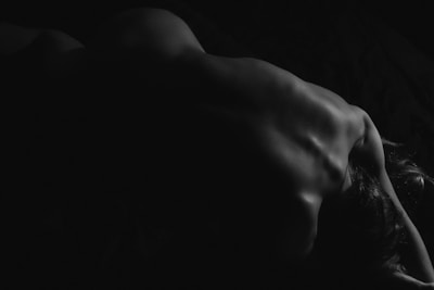 A black and white photograph of a nude back, with a focus on the curvature and shadows that create a dramatic contrast. The light subtly highlights the contours of the spine and shoulder blades.
