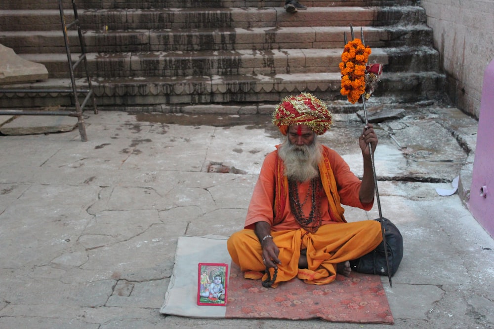 sitting man wearing orange and yellow elbow-sleeved shirt holding staff