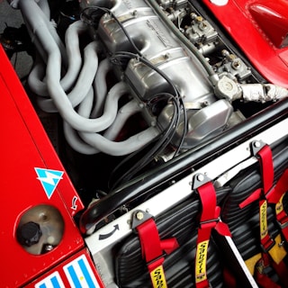 A detailed view of a high-performance car engine with multiple exhaust pipes and various mechanical components. Red safety harnesses are visible, hinting at a racing environment. The engine is housed in a red car with various decals and markings.