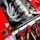 A detailed view of a high-performance car engine with multiple exhaust pipes and various mechanical components. Red safety harnesses are visible, hinting at a racing environment. The engine is housed in a red car with various decals and markings.