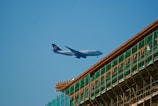 A large passenger airplane with the Lufthansa logo is flying in a clear blue sky. There is a partially constructed or renovated building with scaffolding visible in the foreground.