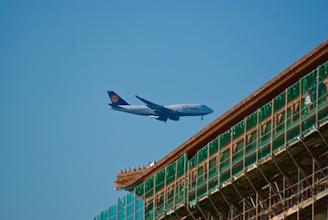 A large passenger airplane with the Lufthansa logo is flying in a clear blue sky. There is a partially constructed or renovated building with scaffolding visible in the foreground.