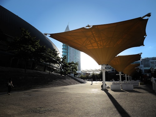 Large architectural canopies create a pattern of shadow and light across a plaza. In the background, modern multi-story buildings are partially illuminated by the sun. A few people can be seen walking through the area, giving a sense of the scale of the structures.