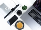 A neatly arranged flat lay featuring modern tech gadgets and accessories. Items include a MacBook, iPhone, Apple Watch, AirPods, a camera lens, a small succulent plant, and a cup of coffee placed on a clean white desk surface alongside a stylish magazine.