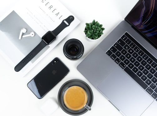 A neatly arranged flat lay featuring modern tech gadgets and accessories. Items include a MacBook, iPhone, Apple Watch, AirPods, a camera lens, a small succulent plant, and a cup of coffee placed on a clean white desk surface alongside a stylish magazine.