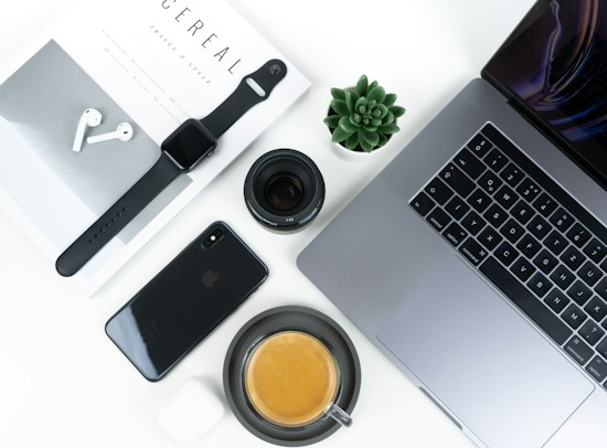 A neatly arranged flat lay featuring modern tech gadgets and accessories. Items include a MacBook, iPhone, Apple Watch, AirPods, a camera lens, a small succulent plant, and a cup of coffee placed on a clean white desk surface alongside a stylish magazine.