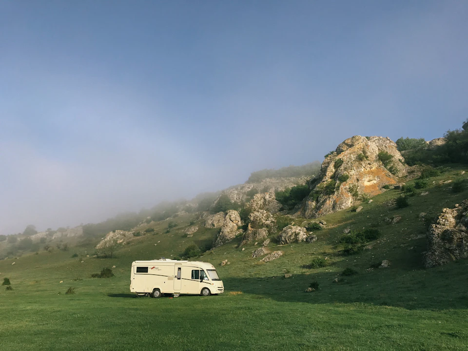 White camper van parked on grassy hillside with lush vegetation and large rocks nearby