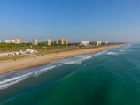 A coastal scene featuring a sandy beach with gentle waves lapping at the shore. Several buildings are visible in the background, indicative of a town or resort area. A few vehicles are parked near the beach, and a couple of people are enjoying the beachfront.