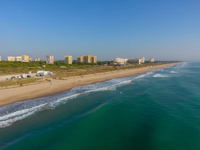 A coastal scene featuring a sandy beach with gentle waves lapping at the shore. Several buildings are visible in the background, indicative of a town or resort area. A few vehicles are parked near the beach, and a couple of people are enjoying the beachfront.