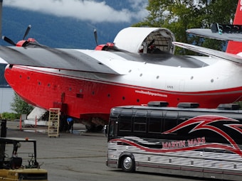 A large red and white aircraft with a distinctive wing design is parked outdoors near a wooded and mountainous area. The aircraft is positioned alongside a large tour bus with the name Martin Mars prominently displayed on the side. Several other vehicles are visible in the vicinity, and there seems to be maintenance or loading activity taking place under the aircraft.