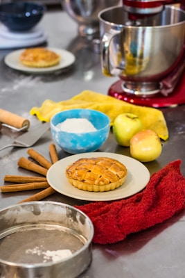 A kitchen scene with a freshly baked lattice pie on a plate, surrounded by apples and cinnamon sticks. A bowl of flour sits next to the pie, along with a rolling pin and a baking tin. A stand mixer is visible in the background, and towels rest on the counter.